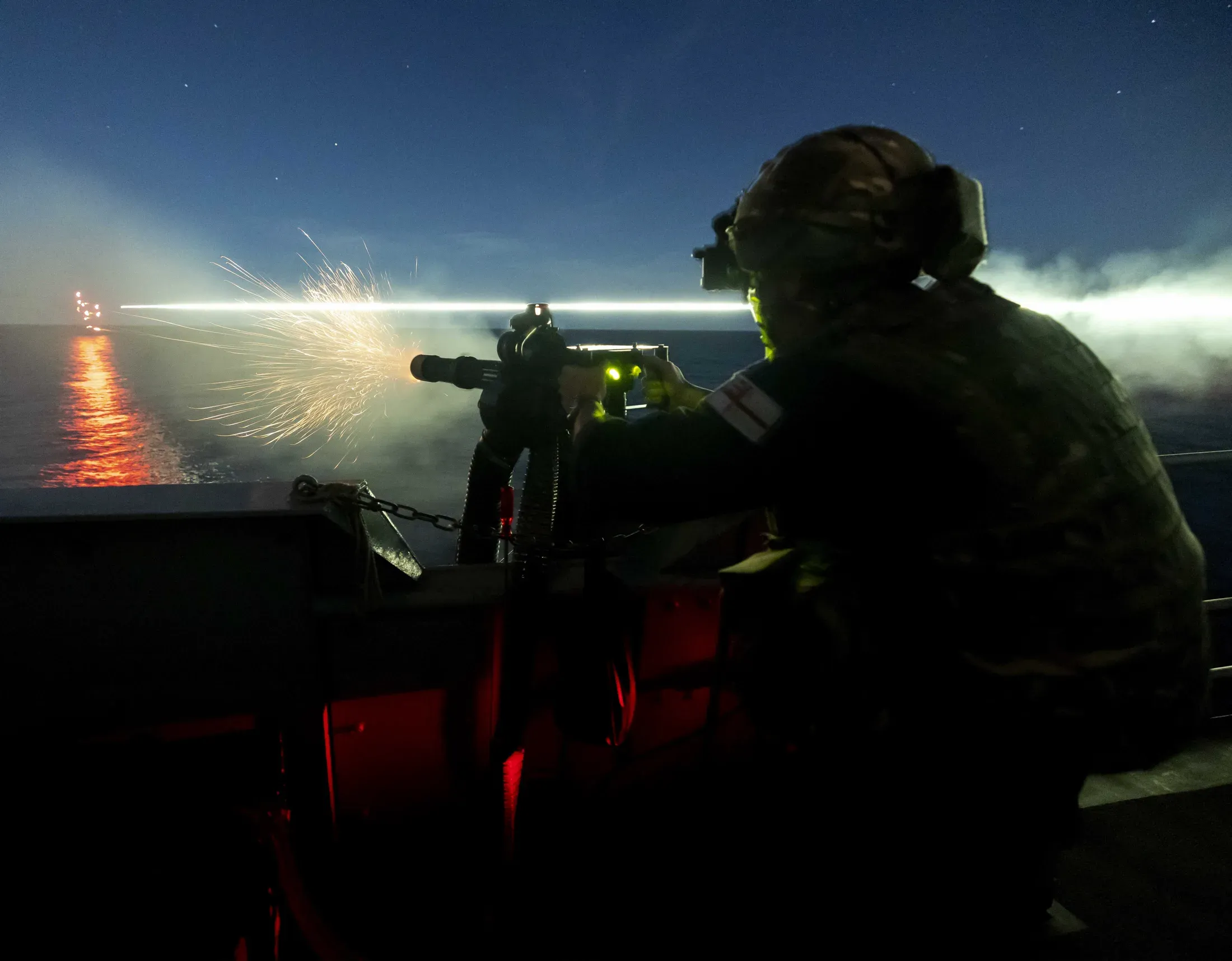 A Royal Navy crew member firing a mounted machine gun at night from a ship at sea